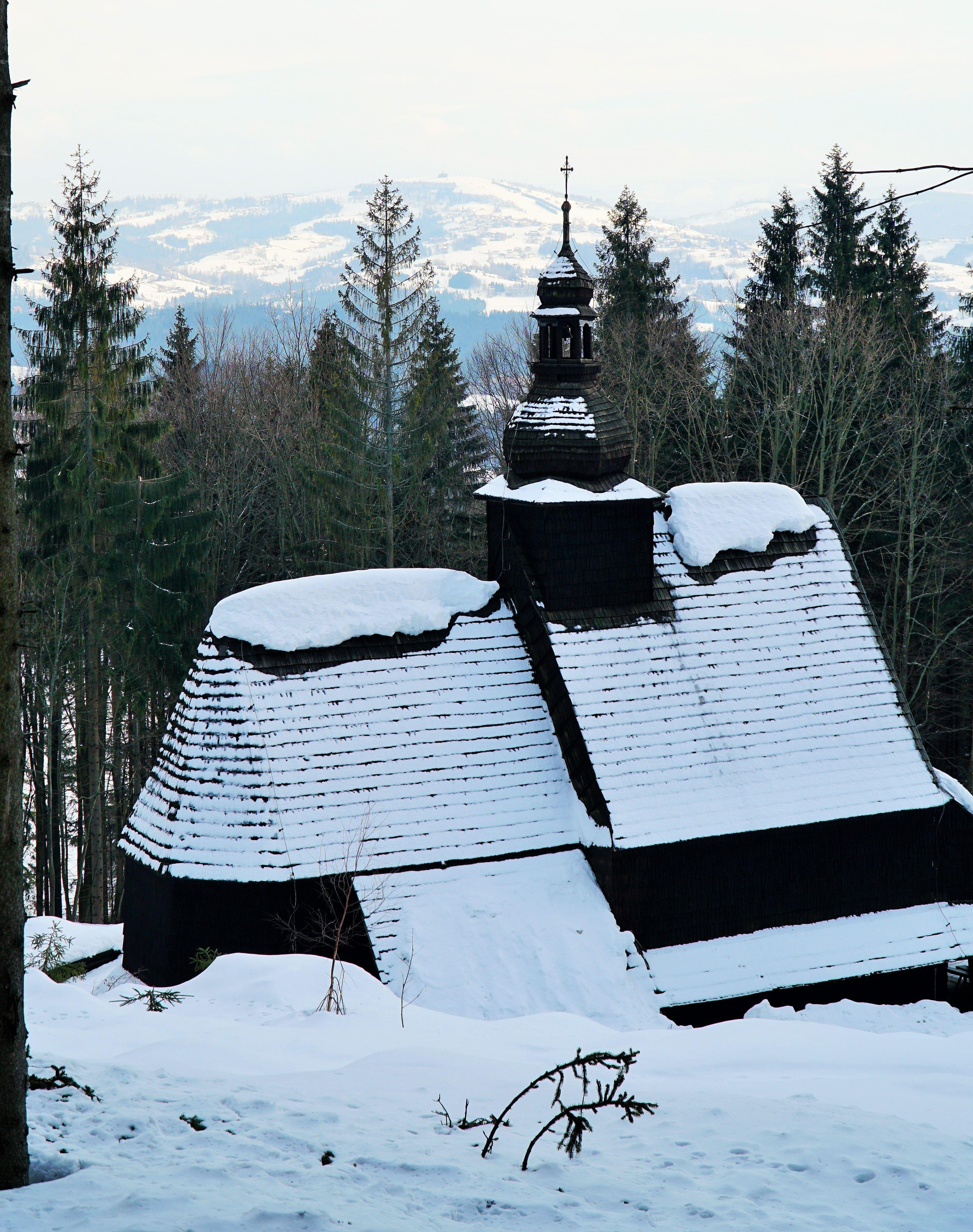 Drewniany kościół widoczny od strony przełęczy w zimowej szacie. W tle drzewa iglaste, na dalekim
planie widok ośnieżonego szczytu Ochodzita w Koniakowie.