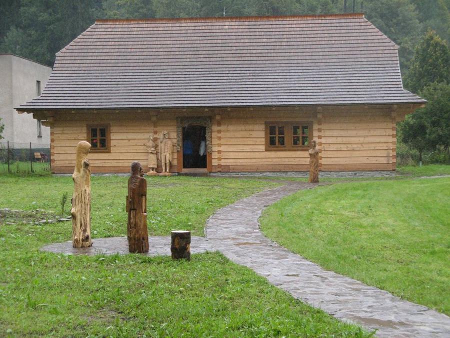 Drewniana chata, do której prowadzi brukowana ścieżka. U wejścia do budynku, jak i wzdłuż ścieżki
znajdują się drewniane rzeźby ludzkich postaci.